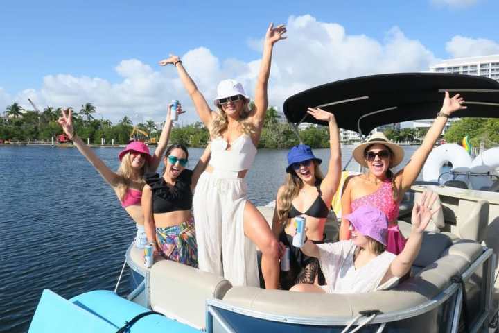 Group of six people on a boat, smiling and posing with arms raised, wearing colorful hats.