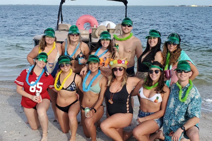 Group of smiling people in swimsuits and hats on a beach near a boat.
